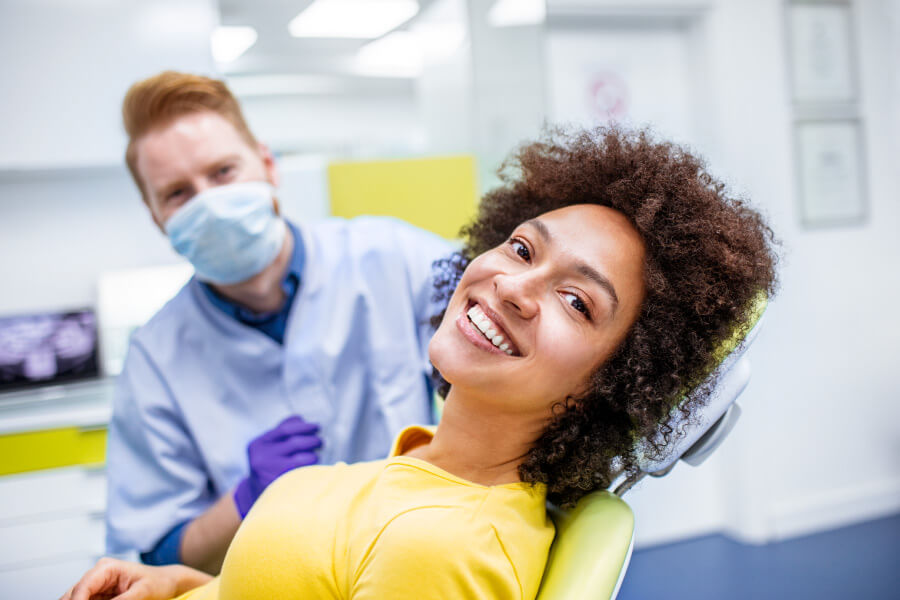 Brunette woman in a yellow shirt smiles while sitting in the dentist chair with a dentist behind her