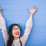Dark-haired woman with a strong immune system smiles and lifts her arms overhead against a blue wall