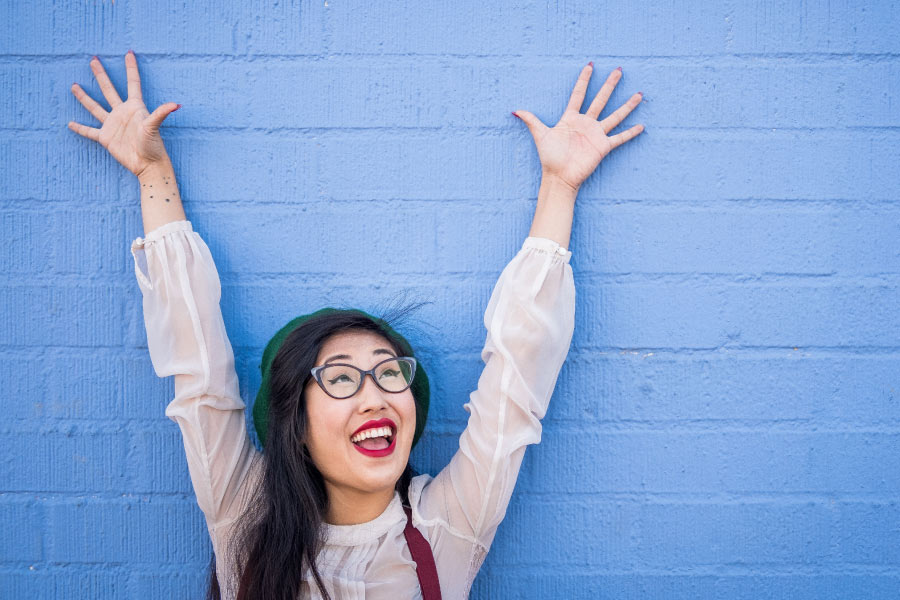 Dark-haired woman with a strong immune system smiles and lifts her arms overhead against a blue wall