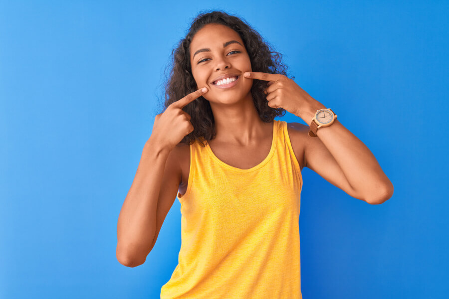 Brunette woman points to her healthy smile while wearing a yellow tanktop against a blue wall